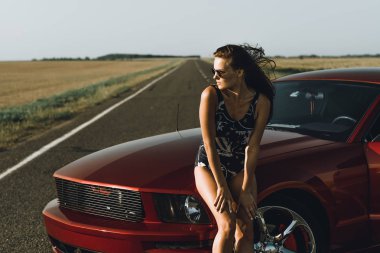 Uzun saçlı ve güneş gözlüğü ile bir bodysuit güzel bir kız yol karşı, kırmızı bir arabanın kaputu üzerinde oturur