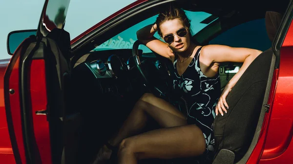 一个穿着长发和太阳镜的紧身衣的漂亮女孩坐在红色汽车的引擎盖上 靠在路上 — 图库照片