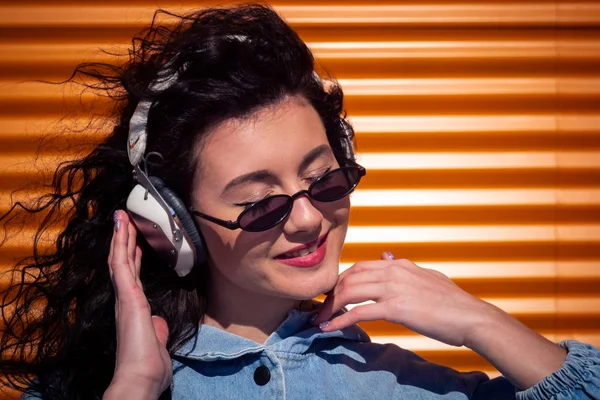 stock image Beautiful young girl with dark curly hair listening to music on headphones, street style, outdoor portrait, hipster girl, music, mp3, Bali, beauty woman, sunglasses, orange color, concept dj