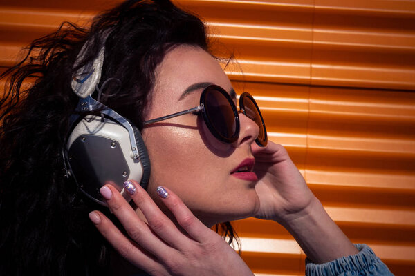 молодая девушка с темными кудрявыми волосами, слушающая музыку в наушниках, уличный стиль, внешний портрет, девушка-хипстер, музыка, mp3, Бали, красавица, солнцезащитные очки, оранжевый цвет, концепт ди-джей
