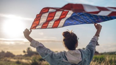 Gözlüklü ve kot pantolonlu güzel bir kadının portresi elinde Amerikan bayrağı sallıyor, mavi gökyüzünün arka planı, vatanseverlik kavramı, gösteri, protesto.