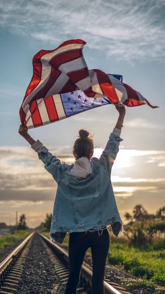 一个戴着眼镜 穿着牛仔裤 手里拿着飘扬的美国国旗 蓝天背景 爱国主义观念 示威和抗议的年轻漂亮女人的画像 — 图库照片