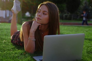 Parkta yeşil çimlerin üzerinde dizüstü bilgisayar kullanan genç bir kadın. Boş zaman aktivite kavramı. 