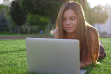 Parkta yeşil çimlerin üzerinde dizüstü bilgisayar kullanan genç bir kadın. Boş zaman aktivite kavramı. 