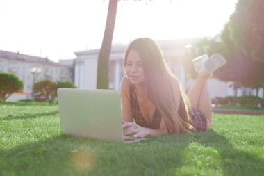 Parkta yeşil çimlerin üzerinde dizüstü bilgisayar kullanan genç bir kadın. Boş zaman aktivite kavramı. 