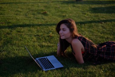 Parkta yeşil çimlerin üzerinde dizüstü bilgisayar kullanan genç bir kadın. Boş zaman aktivite kavramı. 
