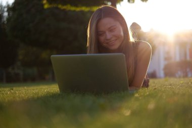 Parkta yeşil çimlerin üzerinde dizüstü bilgisayar kullanan genç bir kadın. Boş zaman aktivite kavramı. 