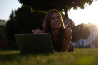 Parkta yeşil çimlerin üzerinde dizüstü bilgisayar kullanan genç bir kadın. Boş zaman aktivite kavramı. 
