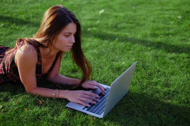 Parkta yeşil çimlerin üzerinde dizüstü bilgisayar kullanan genç bir kadın. Boş zaman aktivite kavramı. 