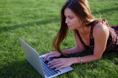 Parkta yeşil çimlerin üzerinde dizüstü bilgisayar kullanan genç bir kadın. Boş zaman aktivite kavramı. 