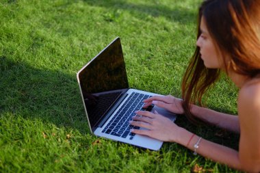 Parkta yeşil çimlerin üzerinde dizüstü bilgisayar kullanan genç bir kadın. Boş zaman aktivite kavramı. 