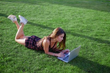 Parkta yeşil çimlerin üzerinde dizüstü bilgisayar kullanan genç bir kadın. Boş zaman aktivite kavramı. 