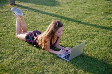 Parkta yeşil çimlerin üzerinde dizüstü bilgisayar kullanan genç bir kadın. Boş zaman aktivite kavramı. 