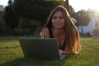 Parkta yeşil çimlerin üzerinde dizüstü bilgisayar kullanan genç bir kadın. Boş zaman aktivite kavramı. 