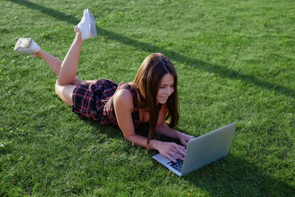 Parkta yeşil çimlerin üzerinde dizüstü bilgisayar kullanan genç bir kadın. Boş zaman aktivite kavramı. 