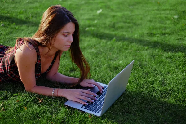 Parkta yeşil çimlerin üzerinde dizüstü bilgisayar kullanan genç bir kadın. Boş zaman aktivite kavramı. 
