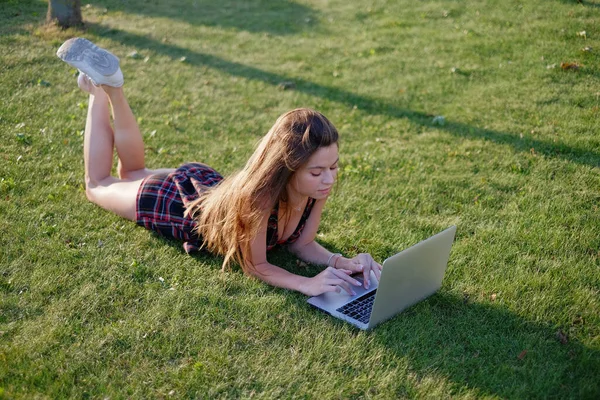 Parkta yeşil çimlerin üzerinde dizüstü bilgisayar kullanan genç bir kadın. Boş zaman aktivite kavramı. 