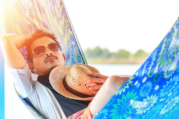 A man wear glasses and sleep on hammock near the river.