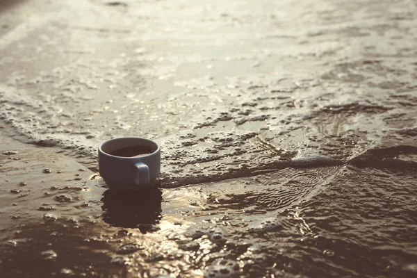 hot cup of coffee on  sand and sea water flow at the beach in the morning.