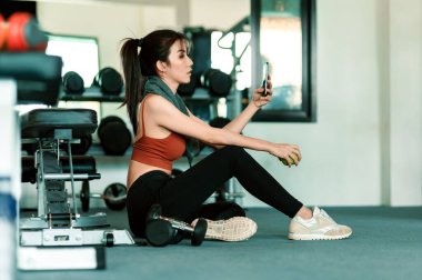 Spor salonunda akıllı telefon kullanan yeşil elmayı tutan bir kız. Spor salonundaki kadın egzersizi ara verip rahatlıyor..