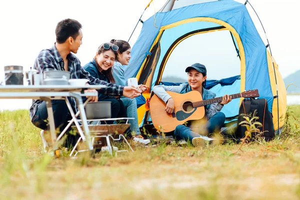 A group of friends camping happily near the lake on a holiday. A woman plays the guitar for a friend to listen