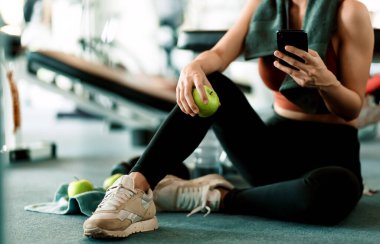 Spor salonunda akıllı telefon kullanan yeşil elmayı tutan bir kız. Spor salonundaki kadın egzersizi ara verip rahatlıyor..