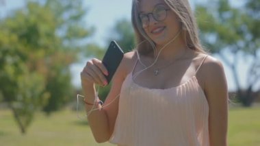 yeşil park ve rüzgar dalgaları yazlık elbise dansları müzik kulaklık üzerinde ağır çekim neşeli sarışın kız bardaklarda dinler