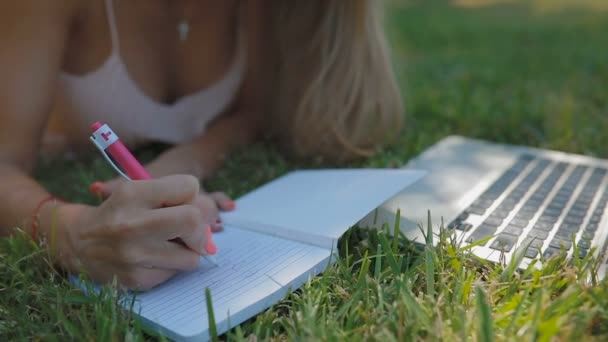 特写镜头的女孩在眼镜与金发碧眼的长而流动的头发躺在草地上画图片与笔在抄本在笔记本电脑 — 图库视频影像