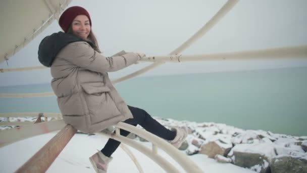 穿着冬衣的慢动作快乐女孩摇着腿坐在雪海滩上方的帐篷管上 举起手来对抗大海 — 图库视频影像