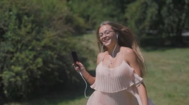 neşeli çekici sarışın kadın ile smartphone üzerinde yeşil yaz park boyunca çalışan kulaklıklar müzik dinler