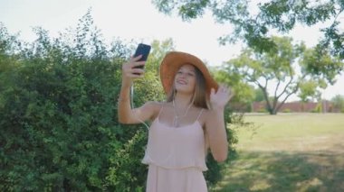 Hasır şapka giyen uzun saçlı kadın parkta telefonda görüntülü konuşma at hava öpücük gönderir