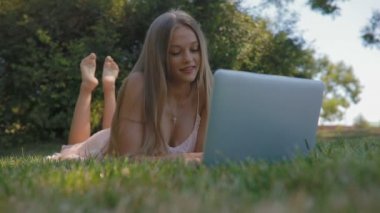çekici sarışın Bayan ile uzun gevşek akan saç üzerinde çim türleri üzerinde Şehir Parkı içinde laptop üzerinde sıcak yaz günü yatıyor