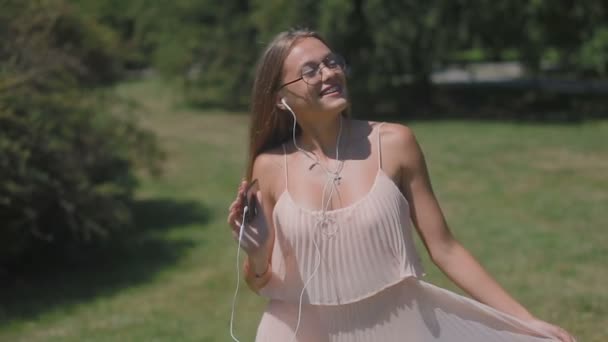スマート フォンでうれしそうな魅力的なブロンドの女性は緑の夏公園に沿って実行しているイヤホンで音楽を聴いてください — ストック動画