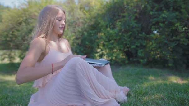 慢动作侧视图金发碧眼的金发女士在粉红色的礼服坐下来 并举行在绿色草地上在城市公园的笔记本电脑 — 图库视频影像