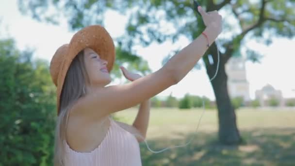 美丽的年轻女子在草帽与手机旋转和波的手在阳光明媚的夏季公园 — 图库视频影像