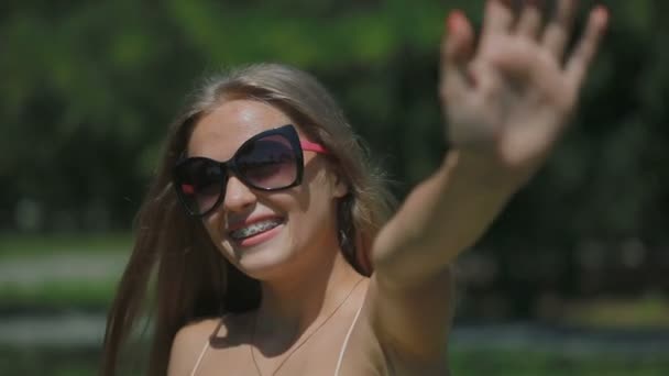 戴着太阳镜的快乐女人微笑着 戴上草帽 挥手挥手到镜头走在公园里 — 图库视频影像