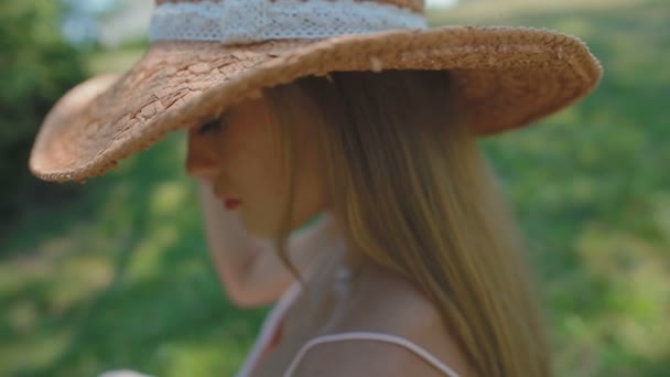 スローモーション クローズ アップ大きな麦藁帽子に魅力的な若い女性が座っているし 明るい日光の下で公園の芝生の上をもたらす — ストック動画