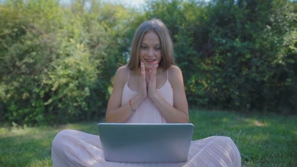 慢动作金发碧眼的女孩 穿着宽松的飘逸的头发 坐在草地上 笔记本电脑拍拍 举起手来以上的头 — 图库视频影像