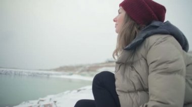 ağır çekim portre dreamy kız kayalık deniz kumsalda oturur ve döner ilham verici ile soğuk ışık kış günü gülümse.