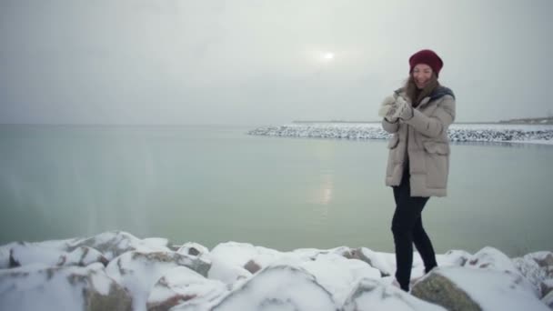 若い幸せな女は 雪をピックアップし 海の近くの岩の上にそれをスロー — ストック動画