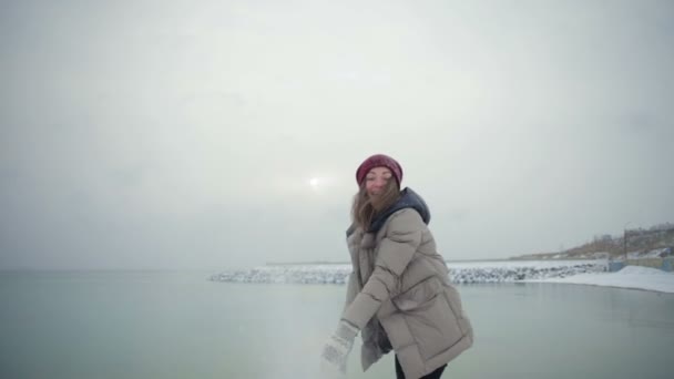 慢动作漂亮的女孩做雪球和投掷以愉快的微笑岩石海滩对苍白的冬日太阳 — 图库视频影像