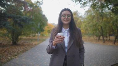 ağır çekim bir palto giyiyor bardaklarda mutlu esmer kız parkta yürüyor ve kağıt bardak kahve içer