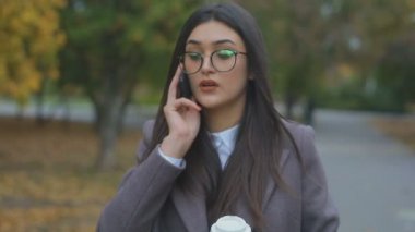 güzel genç kadın smartphone ve kağıt bardak kahve ile sonbahar parkta yürüyor