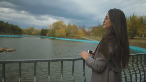 美丽的女孩在时尚的长大衣站在金属扶手反对池塘 — 图库视频影像