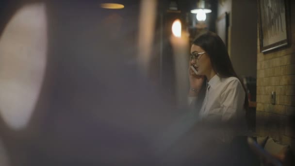 スローモーション眼鏡でぼやけた画像深刻な女性の背後にあるカフェでノート パソコンとテーブルで電話で話す — ストック動画