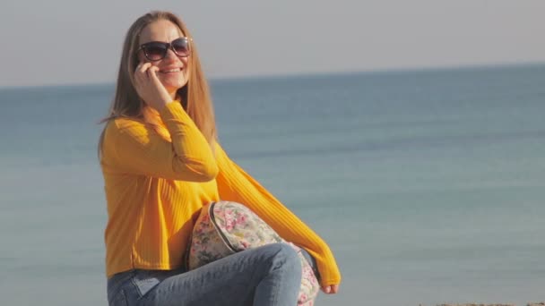 美丽的女人在太阳镜和黄色高领毛衣说话的手机在蓝色的大海在阳光明媚的日子 — 图库视频影像