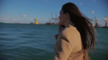 uzun gevşek sallanan saç ile şık esmer mavi deniz yakınında yürür ve liman vinçler yavaş hareket karşı akıllı telefon görüşmeleri