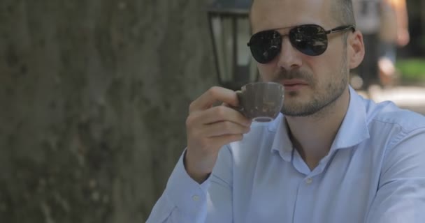 暖かい日に大きな木の影で居心地の良い夏のカフェでホットコーヒーを飲むサングラスの過去の男を閉じる — ストック動画