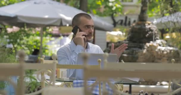 エレガントな男は電話で感情的に話し 噴水と居心地の良い夏のカフェでテーブルから離れて歩きます — ストック動画