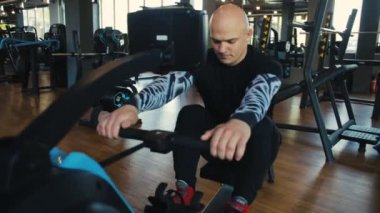Ciddi yakışıklı kel adam kürek makinesine oturur ve boş modern spor salonuna kürek çeker.